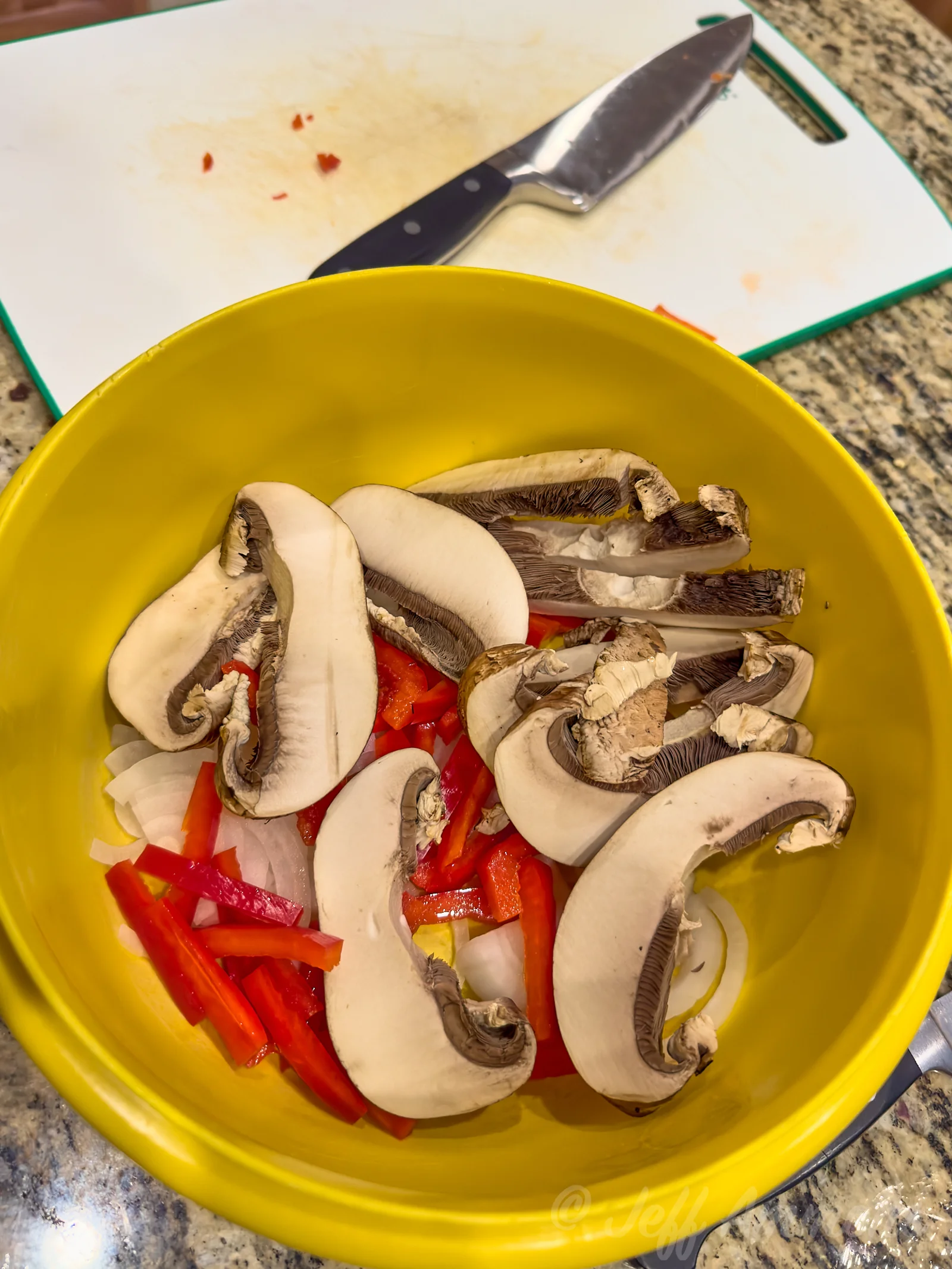 A yellow mixing bowl filled with sliced mushrooms, red bell pepper strips, and chopped onion on a granite countertop, with a cutting board and kitchen knife in the background.
