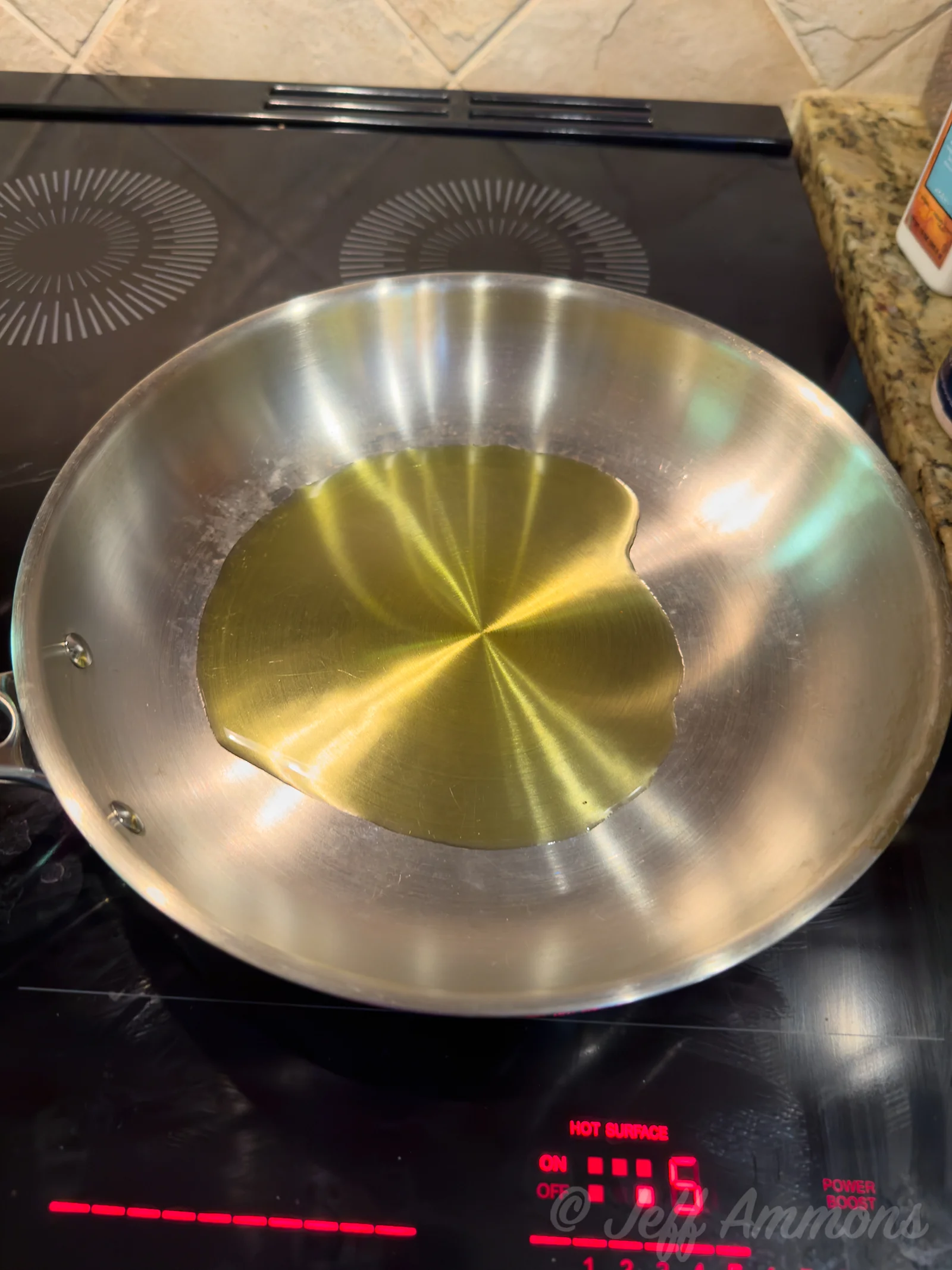 A stainless steel skillet on an induction cooktop with a shallow pool of olive oil warming in the center.