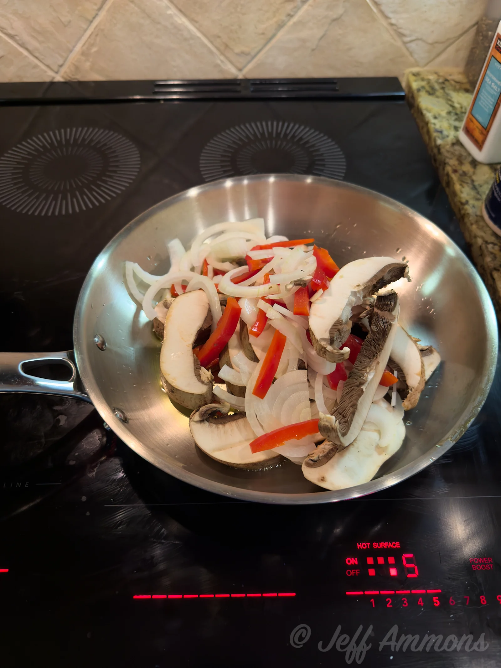 Sliced mushrooms, onion, and red bell pepper strips piled in a stainless steel skillet on an induction cooktop.