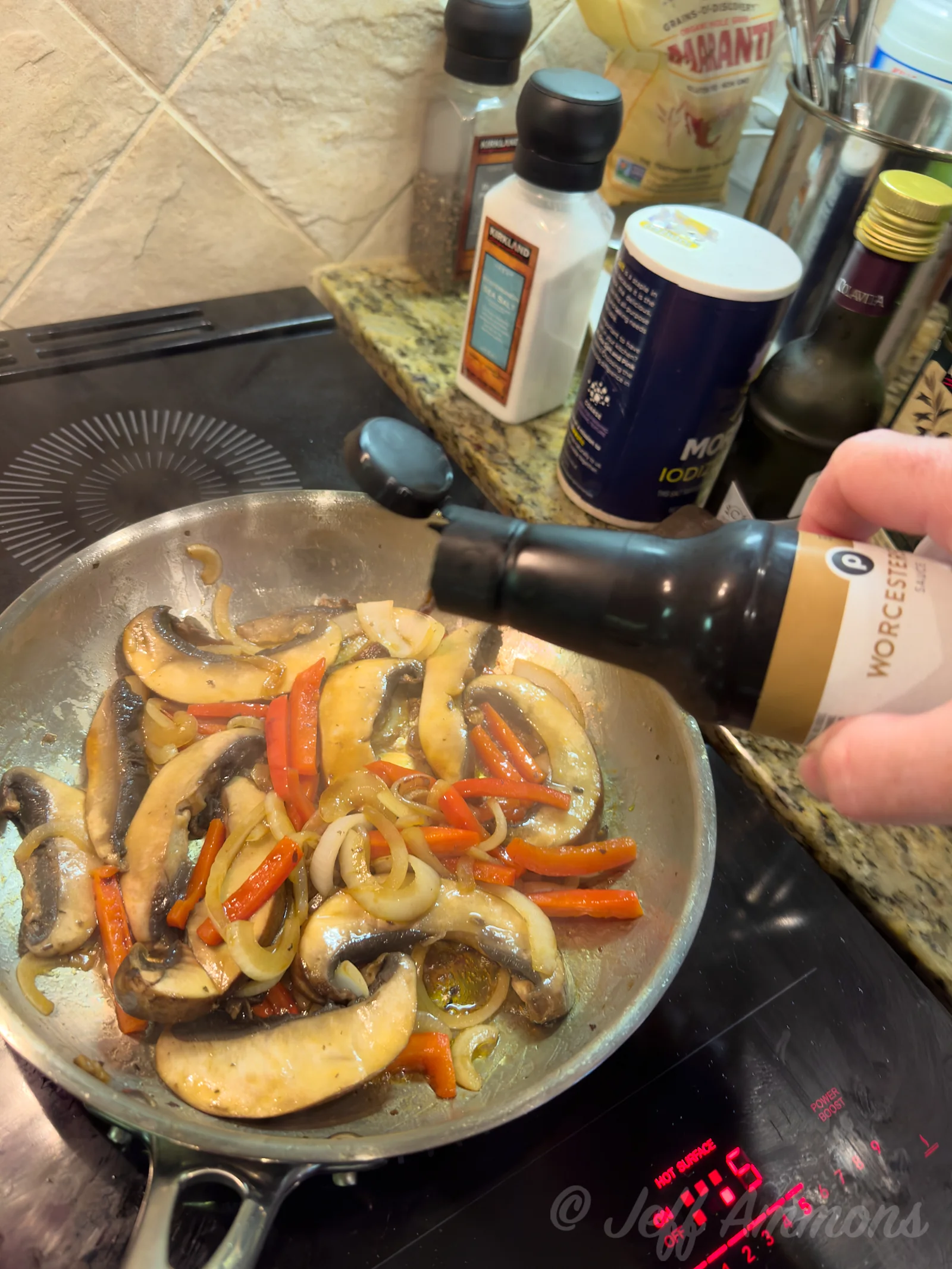 A hand tips Worcestershire sauce into a skillet of sauteed mushrooms, onions, and red bell peppers on the stovetop.