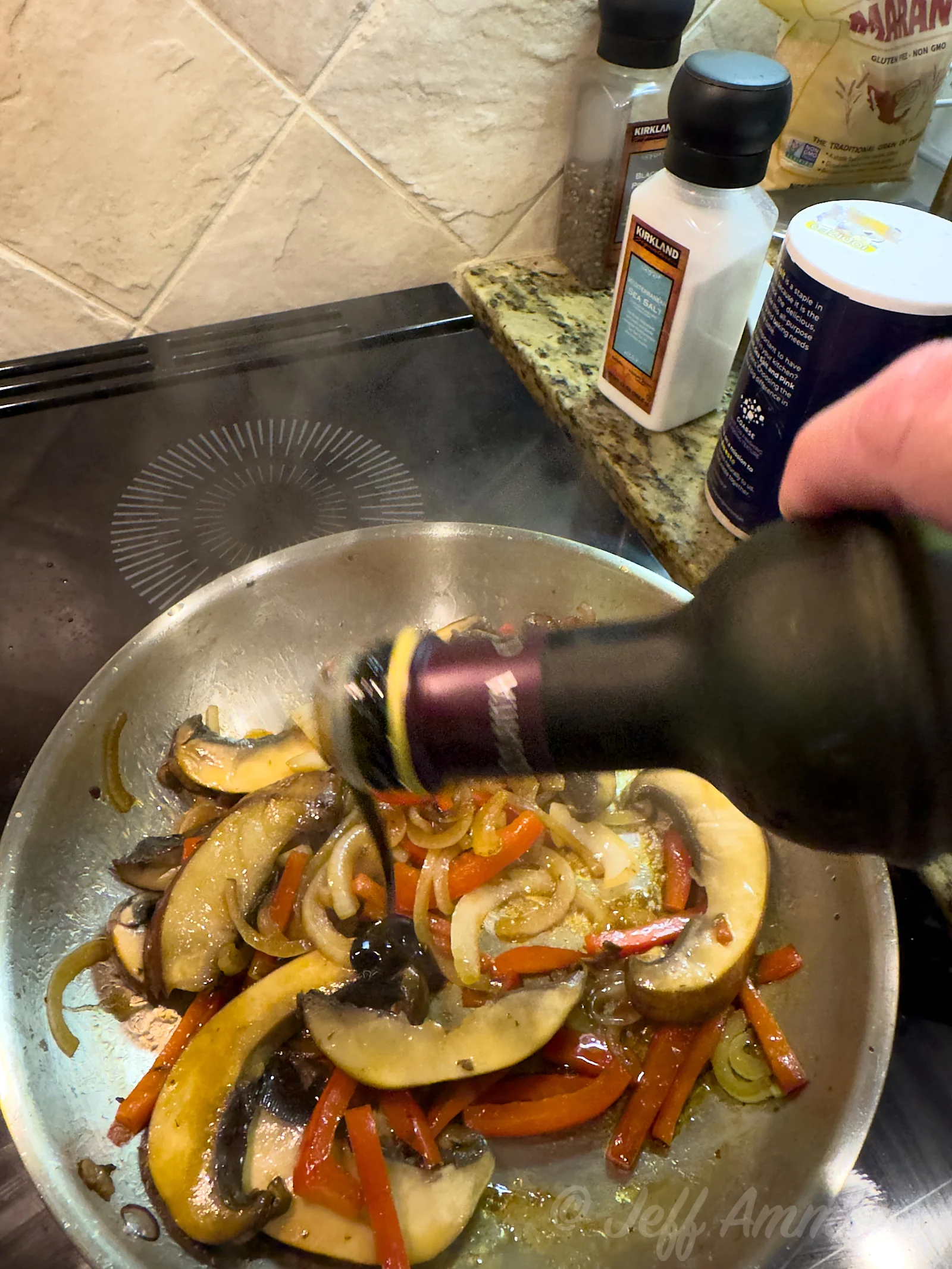 A hand pours a dark balsamic glaze into a skillet of cooked mushrooms, onions, and red bell peppers.