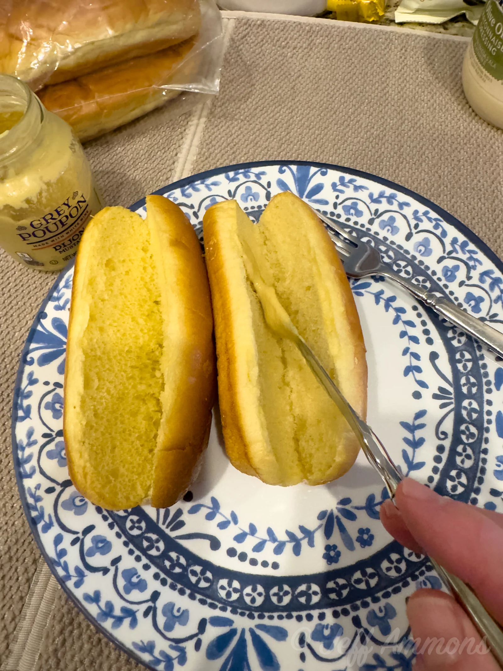 Two split hot dog buns on a blue-and-white plate while a knife spreads Dijon mustard inside one bun.